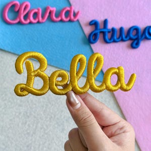 May include: A yellow embroidered patch featuring the name Bella in a cursive script font. The patch has a raised, textured surface with visible thread stitching. In the background, two additional embroidered patches are visible: one pink patch reading Clara and one blue patch reading Hugo.