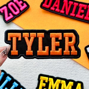 May include: Embroidered patch with the name "TYLER" in orange block letters on a black background. The patch has a black border and is surrounded by other name patches in different colors. The background is orange.