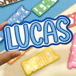 May include: A close-up of a patch with the name "LUCAS" in light blue, outlined in a darker blue, on a white background. Other name patches are visible in the background. The patches are likely for clothing or accessories.