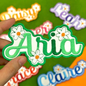 May include: Embroidered name patches in various colors, including green, orange, and blue. The patches feature names like "Aria" and "Claire" in a playful font, decorated with daisy flower accents. The patches are designed for clothing or accessories.