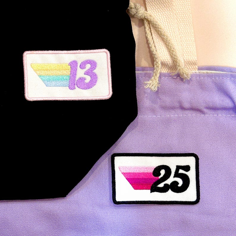 May include: Black and lavender tote bags with embroidered patches. One patch features the number 13 with a rainbow design, and the other displays the number 25 in black with a pink and white design. The bags have a drawstring closure.