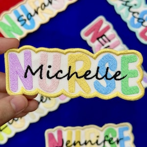 May include: Embroidered patch with the word "Nurse" in pastel colors, with the name "Michelle" in black script. The patch has a yellow border and is on a white background. Other patches with names are visible.