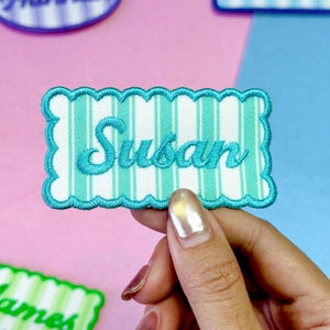 May include: A rectangular embroidered patch with a scalloped edge in bright teal. The patch features a white and teal vertical striped pattern with the name Susan stitched in teal cursive script across the center of the fabric surface.