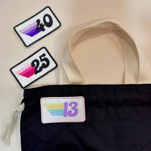 May include: A black tote bag with a cream handle and three rectangular patches. The patches have a white background, black borders, and the numbers 40, 25, and 13. The patches feature gradient colour schemes.