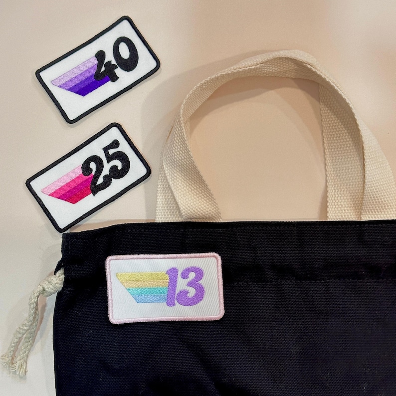May include: A black tote bag with a cream handle and three rectangular patches. The patches have a white background, black borders, and the numbers 40, 25, and 13. The patches feature gradient colour schemes.