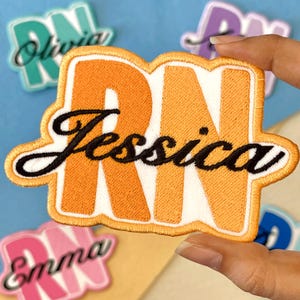 May include: An orange and white embroidered patch with the letters "RN" and the name "Jessica" in black script. The patch has a white border and is held up against a blue background with other patches.