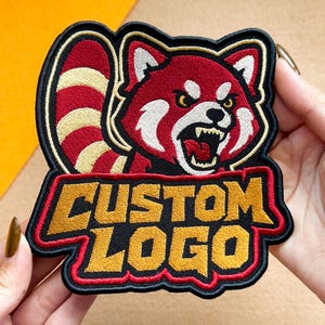 May include: An embroidered patch featuring a red panda with a striped tail and an aggressive expression, with its mouth open showing teeth. Below the red panda, the text CUSTOM LOGO is stitched in bold yellow letters with a red border on a black background.