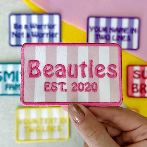 May include: A rectangular embroidered patch featuring a light pink and white vertical striped background with a dark pink stitched border. The text "Beauties" is embroidered in dark pink, followed by the text "EST. 2020" in the same color.