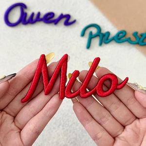 May include: A red embroidered patch featuring the name Milo in a cursive script font. The patch is held up against a light background, with additional embroidered name patches in purple and teal visible in the blurred background area.