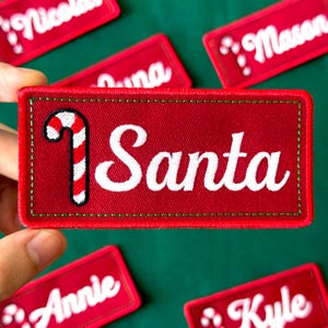 May include: Red fabric patch with the word "Santa" in white script, a candy cane graphic, and a green border. Other name patches are visible. Festive holiday decor.