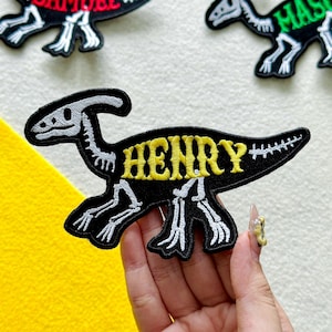 May include: An embroidered iron-on patch shaped like a dinosaur skeleton in black and white thread. The dinosaur features the name HENRY stitched in bold yellow letters across its body. The design includes a long tail and a crested head shape.