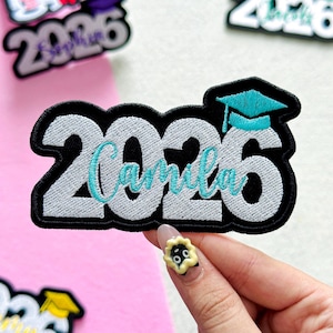 May include: Embroidered patch with the year "2026" in white, outlined in black, and the name "Camila" in teal script. A teal graduation cap sits atop the "6". The patch is on a pink and white background.