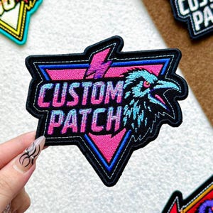 May include: Embroidered patch with the words "CUSTOM PATCH" in a gradient pink and blue font. The patch features a pink triangle with a blue outline and a black raven design. A pink lightning bolt is above the text.