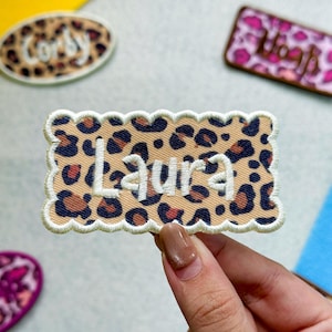 May include: A rectangular embroidered patch with a scalloped edge featuring a tan, brown, and black leopard print pattern. The name Laura is stitched in white thread across the center of the patch. The patch has a white border.