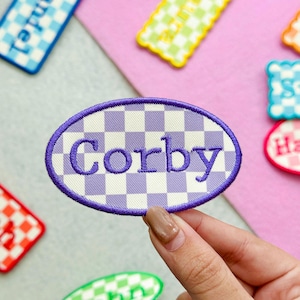 May include: An oval-shaped embroidered patch featuring a light purple and white checkered pattern. The name Corby is stitched in the center in a bold, dark purple font. The patch is finished with a dark purple stitched border edge.