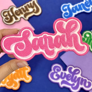 May include: Embroidered patch in a cursive font spelling "Sarah" in bright pink with a white outline. Other name patches in various colors are visible in the background. The patches are likely for clothing or accessories.