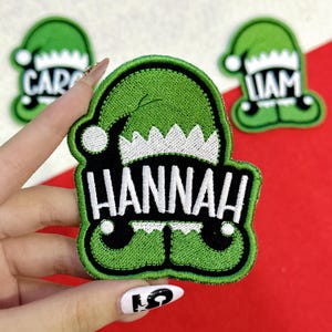 May include: Green embroidered patch in the shape of a Christmas elf with the name "HANNAH" in white block letters. The patch has a black border and white accents. Two other similar patches with names are in the background.
