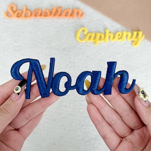 May include: A dark blue embroidered name patch featuring the name Noah in a cursive script font. The patch is held up against a light background where other name patches with the text Sebastian and Capheny are visible in the distance.
