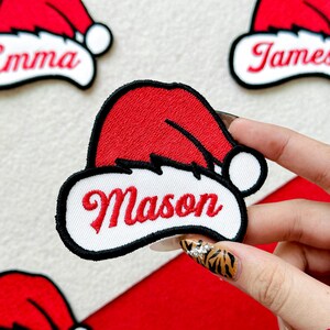 May include: Embroidered red Santa hat patch with the name "Mason" in red script. The patch has a white border and a black outline. Additional patches with names are visible.