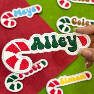May include: Candy cane-shaped patches with names embroidered on them. The patch held in the hand says "Alley" in green letters. Other patches say "Mayer", "Cole", "Simon", and "Serer". The candy canes are red and white.