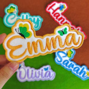 May include: Embroidered name patches with butterfly accents. The patches feature names like "Emma", "Hannah", "Sarah", "Olivia", and "Emily" in various colors. Each patch has a white background with colorful embroidery.