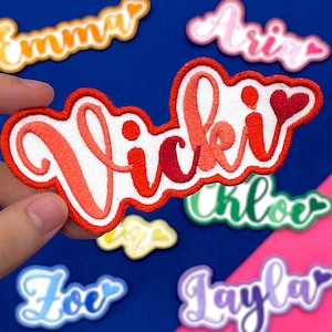 May include: Embroidered name patches in various colours. The patch held in the image says "Vicki" in red and orange thread with a white background and a red border. Other patches with names like "Emma", "Aria", "Chloe", "Zoe", and "Layla" are also visible.