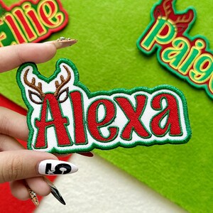 May include: Embroidered patch with the name "Alexa" in red letters with a white border and green outline. The patch features brown antlers above the name and is held against a green felt background. Other patches are visible.
