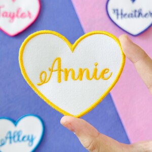 May include: A white heart-shaped patch with a yellow embroidered border and the name "Annie" in yellow script. The patch is held by a hand, with other heart-shaped patches visible in the background.