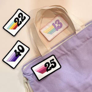 May include: Four embroidered patches with numbers and rainbow or gradient designs. The numbers are 22, 13, 40, and 25. A lilac tote bag with a cream-coloured handle is in the background.
