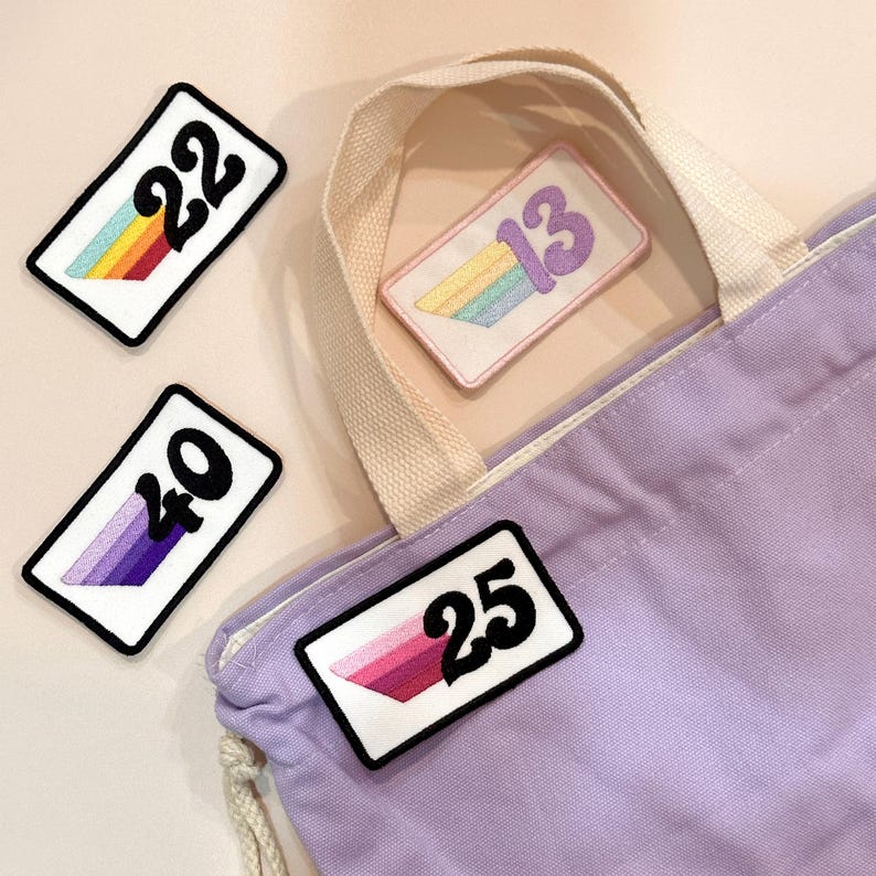 May include: Four embroidered patches with numbers and rainbow or gradient designs. The numbers are 22, 13, 40, and 25. A lilac tote bag with a cream-coloured handle is in the background.