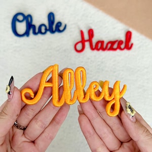 May include: Embroidered name patches in blue, red, and orange. The orange patch spells "Alley" in a cursive font. The blue patch spells "Chloe" and the red patch spells "Hazel". These patches are likely for clothing or accessories.