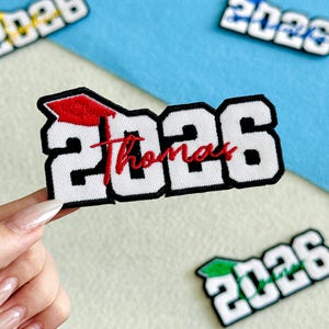 May include: White and black embroidered patch with the text "2026 Thomas" and a red graduation cap. The patch is held up against a blue and white background. Other patches with the year 2026 are visible.