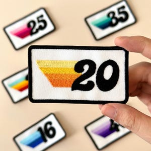 May include: Rectangular white embroidered patches with black borders and numbers. The central patch displays the number 20 in black, with a retro-style design in yellow, orange, and gold. Other patches show numbers 16, 25, and 35.