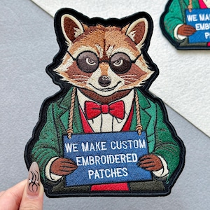 May include: Embroidered patch of a raccoon wearing glasses, a red bow tie, and a green jacket. The raccoon holds a blue sign with the text "WE MAKE CUSTOM EMBROIDERED PATCHES". The patch has a black border.