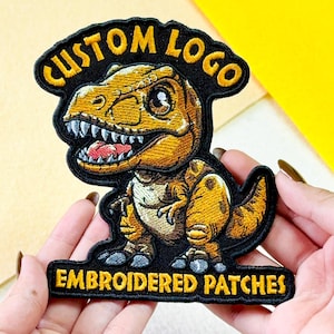 May include: An embroidered patch featuring a cartoon Tyrannosaurus Rex in shades of yellow, brown, and white. The text CUSTOM LOGO is arched above the dinosaur, and EMBROIDERED PATCHES is printed in yellow on a black base at the bottom of the patch.