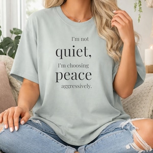 Könnte beinhalten: Ein helles salbeigrünes Kurzarm-T-Shirt mit dem schwarzen Aufdruck "I'm not quiet, I'm choosing peace aggressively." Das Shirt hat einen Rundhalsausschnitt und eine lockere Passform und besteht aus einfarbigem Stoff mit matter Oberfläche.