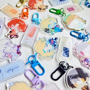 Puede incluir: Una colección de llaveros de acrílico transparente con ilustraciones de personajes de estilo anime y texto. Los llaveros tienen cierres de diferentes colores, incluyendo dorado, naranja, verde, azul, morado y negro. Algunos llaveros tienen la palabra "HELLO!!".