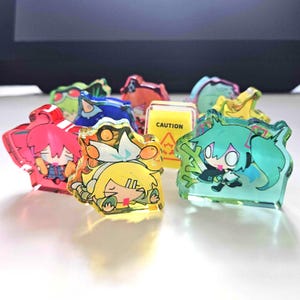 Puede incluir: Una colección de coloridos amuletos de acrílico transparente con personajes de estilo anime. Los amuletos son de varios colores, incluyendo rosa, amarillo, verde y azul. Un amuleto tiene un letrero de precaución amarillo. Los personajes tienen ojos grandes y rasgos estilizados.