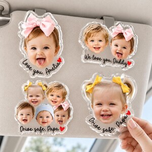 May include: Clear acrylic car visor charms featuring smiling baby faces with bows. Text includes "Love you, Amelia", "We love you, Daddy", and "Drive safe, Papa", with heart accents. A personalized gift.