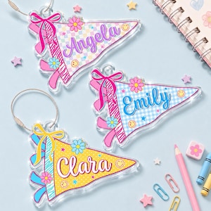 May include: Three triangular acrylic keychains featuring personalized names Angela, Emily, and Clara. Each keychain has a colorful patterned background, a decorative bow, small flower and smiley face icons, and a silver metal wire ring attachment.