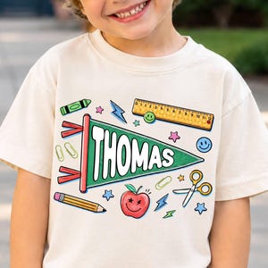 May include: A cream-colored t-shirt featuring a graphic print of a green pennant flag with the name THOMAS in white letters. The design includes school-themed illustrations of a yellow ruler, a pencil, scissors, an apple, and paper clips.