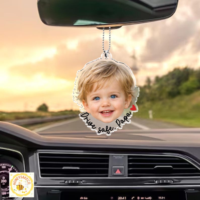 May include: A clear, circular car air freshener with a smiling child's face and the words "Drive safe, Papa." The image has a small red heart and hangs from a silver chain. The background shows a car interior and a road.