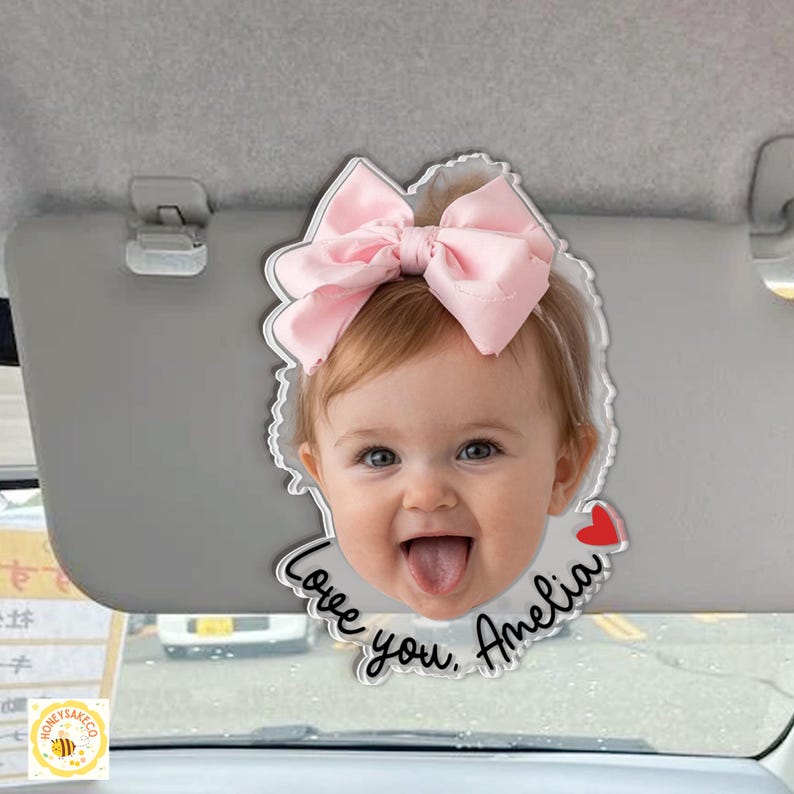 May include: A clear acrylic car visor ornament featuring a smiling baby with a pink bow. The ornament has the text "Love you, Amelia" and a small red heart. The ornament is hanging from a car visor.