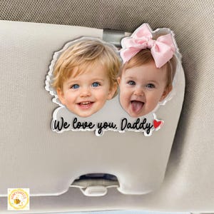 May include: A clear acrylic car visor clip featuring two smiling babies with blue eyes. The clip has the text "We love you, Daddy" with a red heart. One baby wears a pink bow.