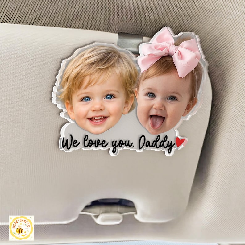 May include: A clear acrylic car visor clip featuring two smiling babies with blue eyes. The clip has the text "We love you, Daddy" with a red heart. One baby wears a pink bow.