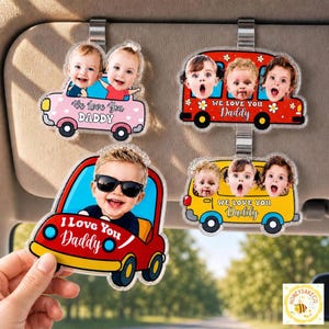 May include: Four car visor clips with photos of babies and children. The clips are shaped like cars and buses in pink, red, and yellow. Each clip has the text "We Love You Daddy" or "I Love You Daddy".