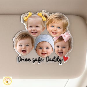 May include: A clear acrylic car visor decoration featuring five smiling baby faces. The text "Drive safe. Daddy" is written in a cursive font, with a red heart at the end. The babies have various hair colors and wear bows and a striped hat.