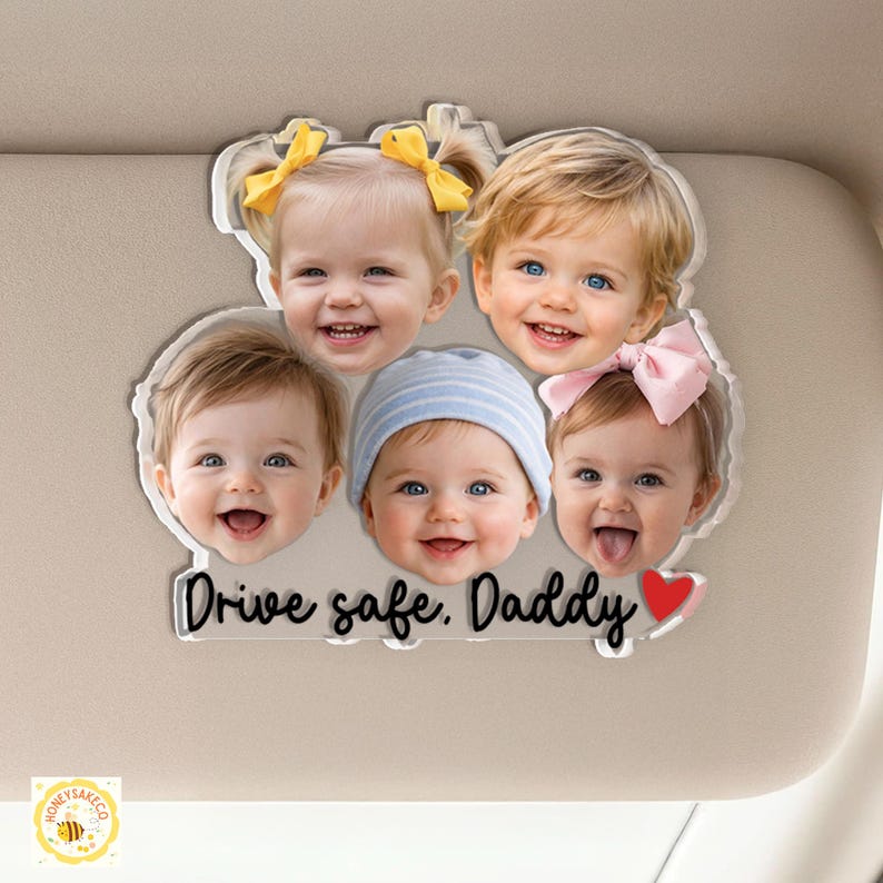 May include: A clear acrylic car visor decoration featuring five smiling baby faces. The text "Drive safe. Daddy" is written in a cursive font, with a red heart at the end. The babies have various hair colors and wear bows and a striped hat.