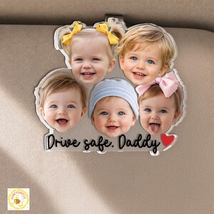 May include: A clear acrylic car visor clip featuring the smiling faces of five babies. The text "Drive safe, Daddy" is written in a cursive font, with a red heart. The clip is designed to attach to a car visor.