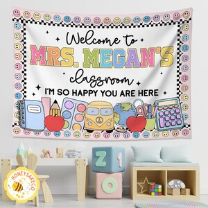 K&ouml;nnte beinhalten: Ein Wandteppich f&uuml;r das Klassenzimmer mit dem Text "Welcome to MRS. MEGAN'S classroom. I'M SO HAPPY YOU ARE HERE." Das Design umfasst Illustrationen zum Thema Schule wie einen Apfel, einen Globus und einen Bus, umrahmt von einem Rand aus Smileys und Schachbrettmustern.